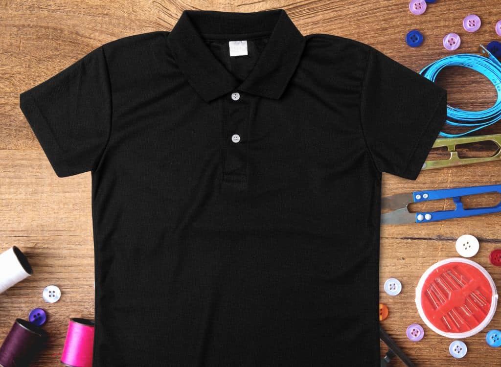 Black custom polo shirt on a wooden surface surrounded by sewing and tailoring accessories, including buttons, thread spools, scissors, and measuring tape.