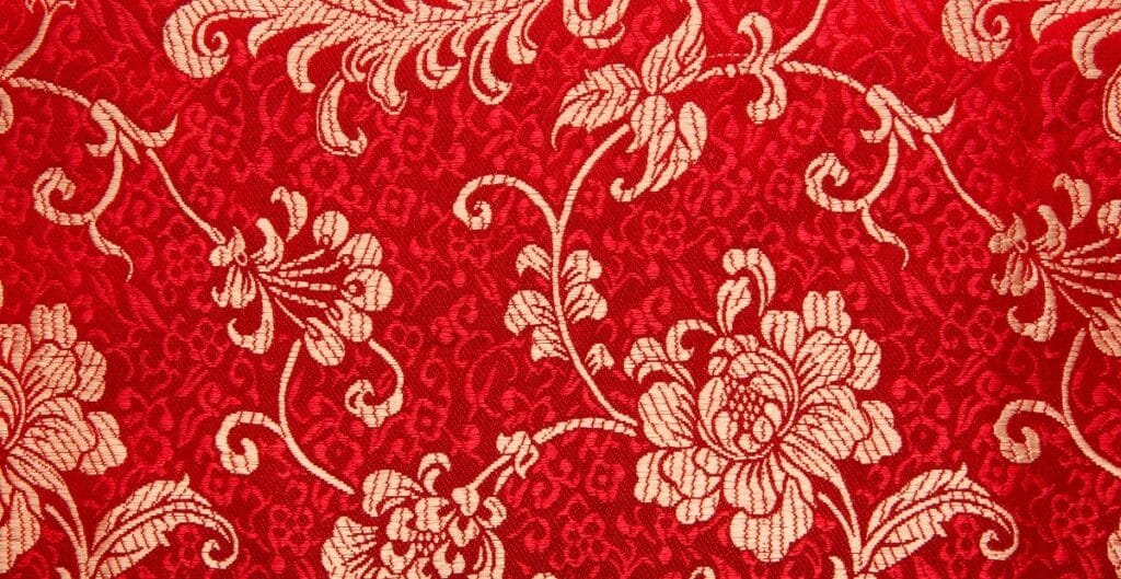 Close-up image of a red designer cotton fabric with an intricate cream floral pattern.