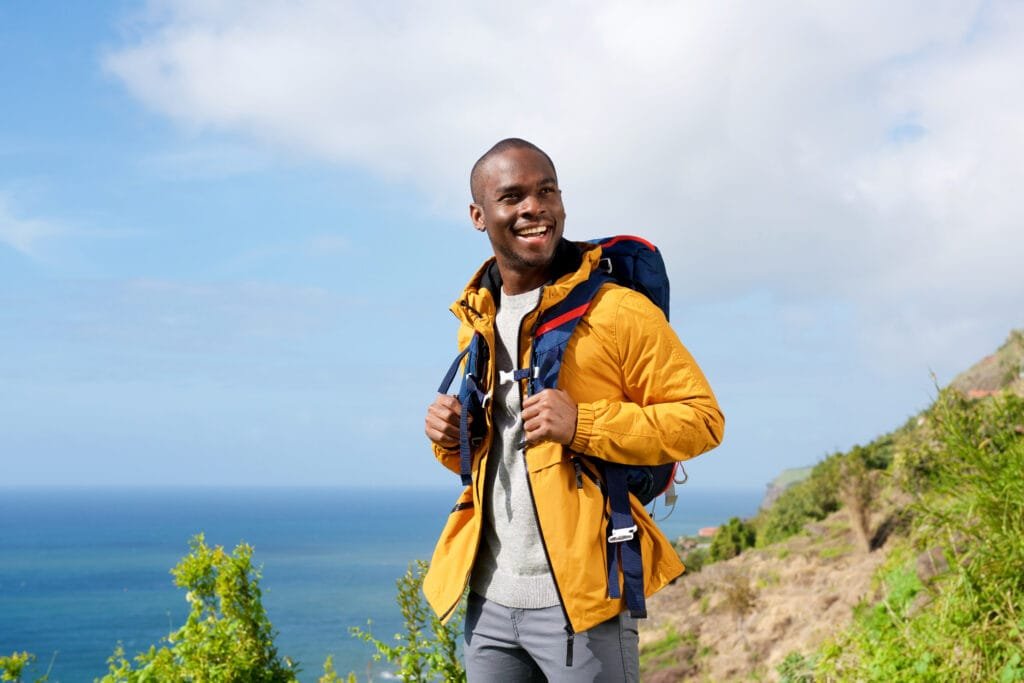  A smiling man wearing a yellow Columbia sports jacket and carrying a backpack, standing on a scenic outdoor trail with a view of the ocean and hillside