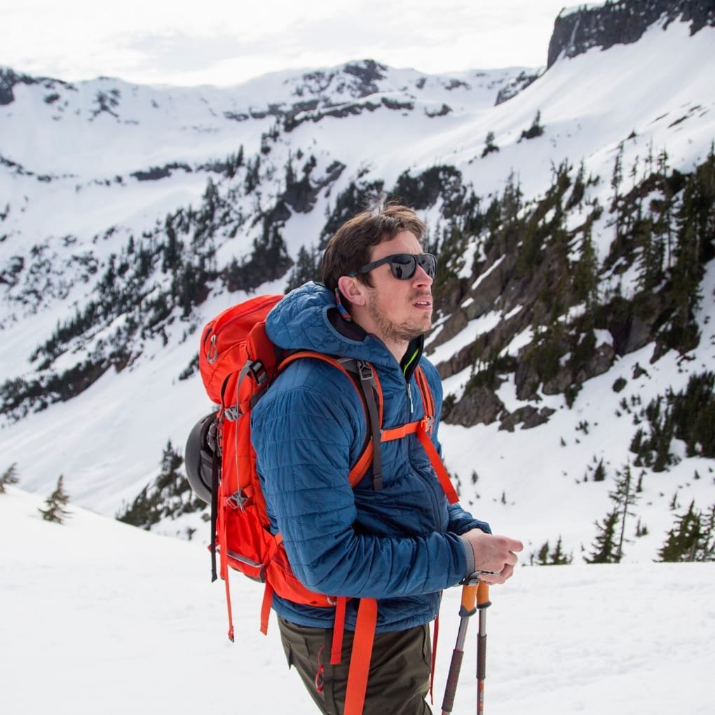 A man dressed in a blue jacket with a red backpack and sunglasses, hiking in a snowy mountain landscape with trees and snow-covered peaks.