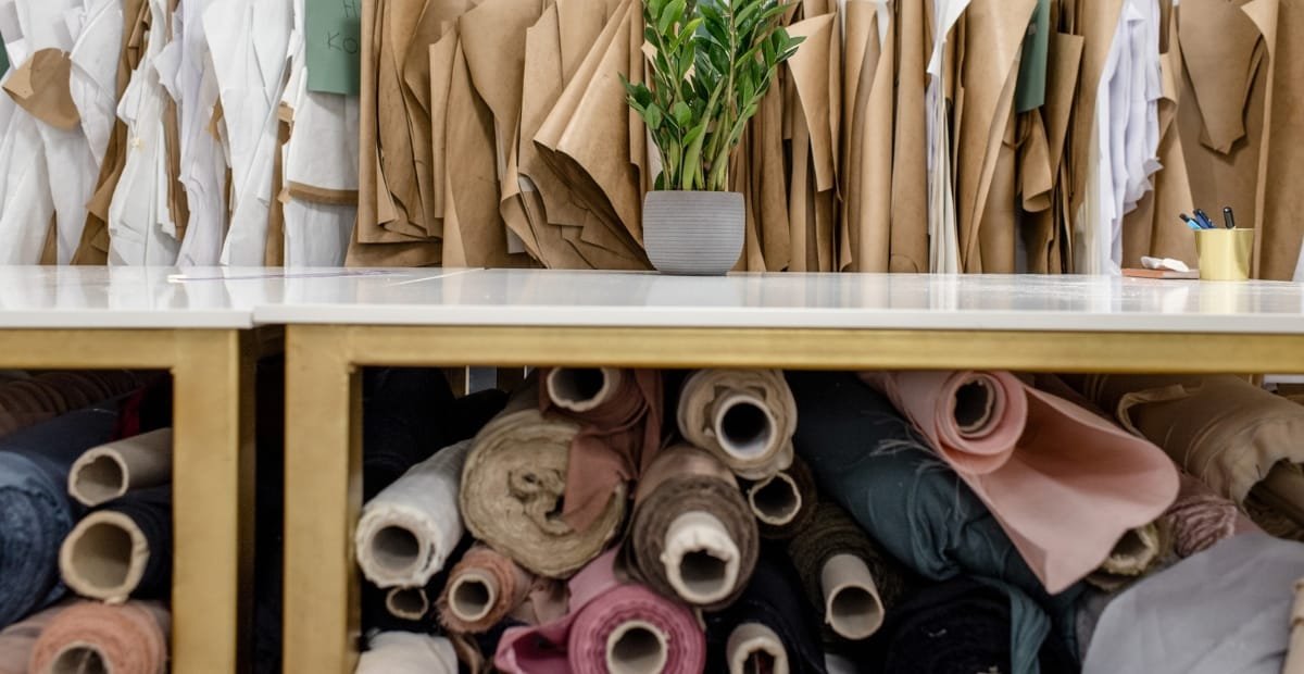 A storage area with a table and fabric rolls underneath it. The table has a potted plant, a mug with pens, and some paper clips. Behind the table, there are neatly arranged brown and white fabric bundles hanging vertically, and below, rolls of fabric in various colors are stored.