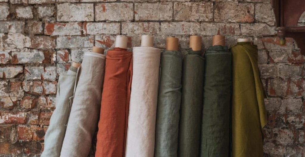 Several rolls of fabric in neutral and earthy tones hanging against a rustic brick wall.