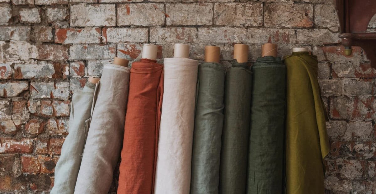 Several rolls of fabric in neutral and earthy tones hanging against a rustic brick wall.