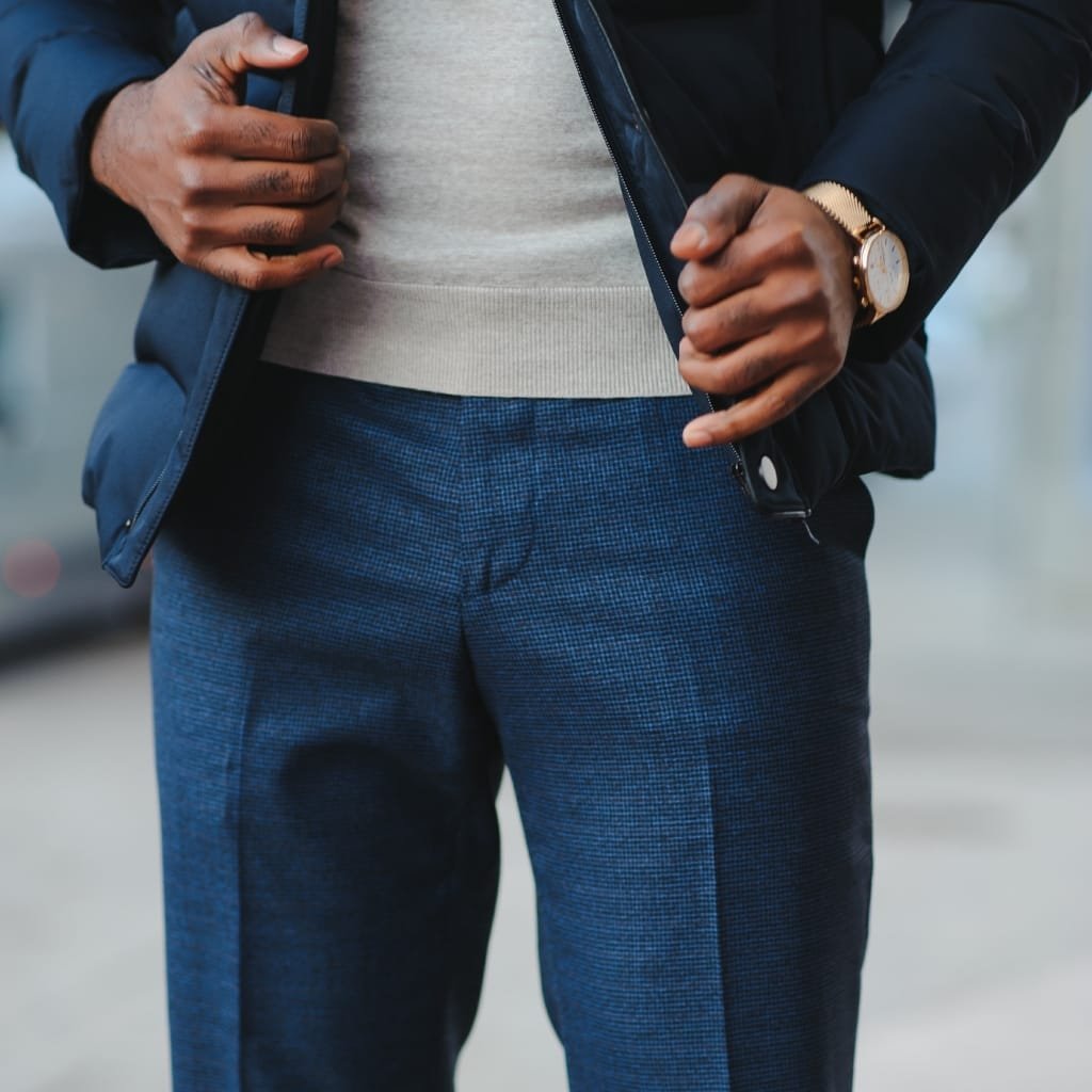 Close-up of a person wearing chambray blue pants, with hands adjusting a dark jacket over a light grey sweater, and a gold wristwatch on their left wrist.