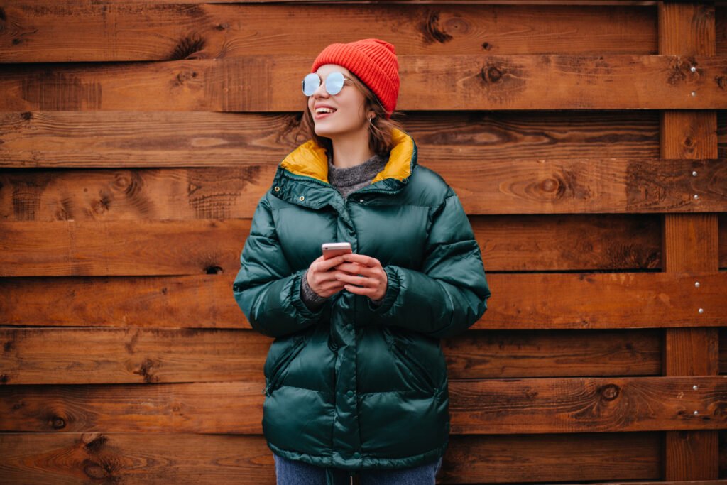 A woman wearing a teal Columbia insulated coat with a yellow lining, standing against a wooden fence, holding a phone, and smiling with sunglasses and a red knit hat.