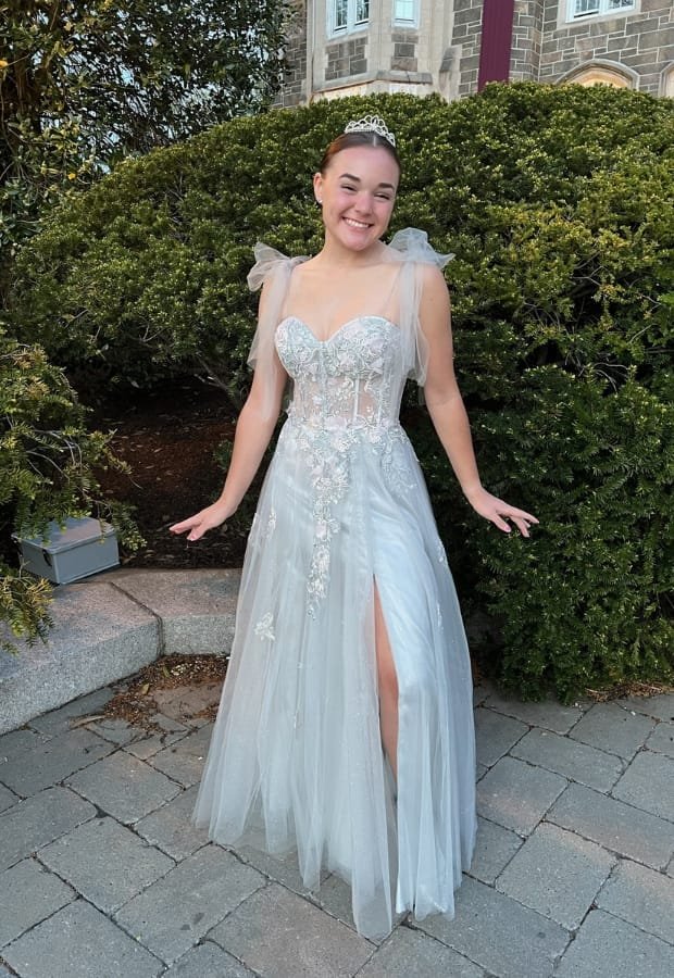 A young woman in a light coral prom dress with floral embellishments, standing outdoors on a paved area with greenery and a stone building in the background, smiling and posing.