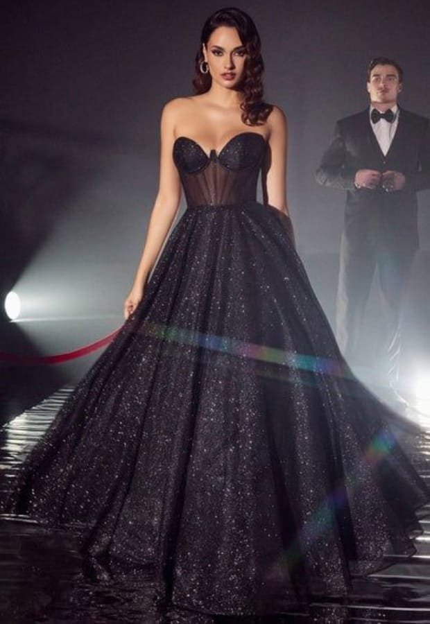 A woman in a glamorous black prom dress with lace and glittery details, standing on a runway or stage, with a man in a tuxedo in the background.