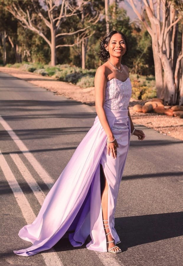 A young woman in a lavender prom dress standing on a road lined with trees, smiling and looking cheerful.