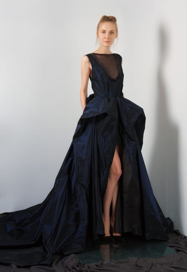 A young woman wearing a striking midnight blue prom dress with a high slit, standing against a plain background. The dress features a sheer black neckline and an elaborate, voluminous skirt with a train.