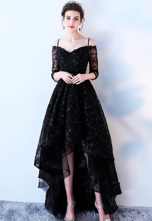 A woman wearing a stunning black prom dress with a high-low hemline, featuring lace details and off-shoulder design, paired with silver high heels.