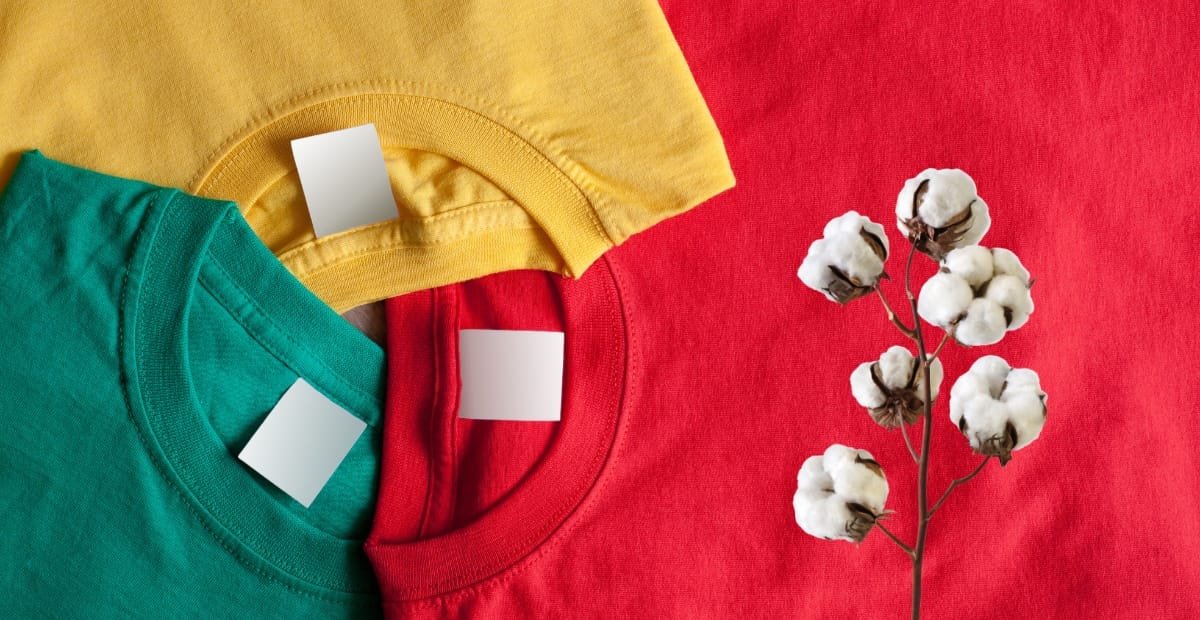 Three colorful T-shirts (yellow, green, and red) are folded and stacked with blank white tags, placed next to a cotton branch on a red fabric background.