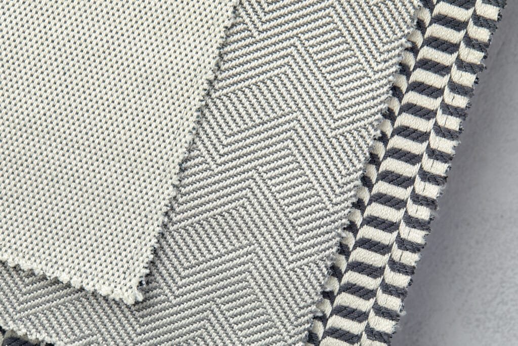 Close-up image of layered textured fabrics featuring different weaving patterns, including herringbone and striped designs, in neutral tones.