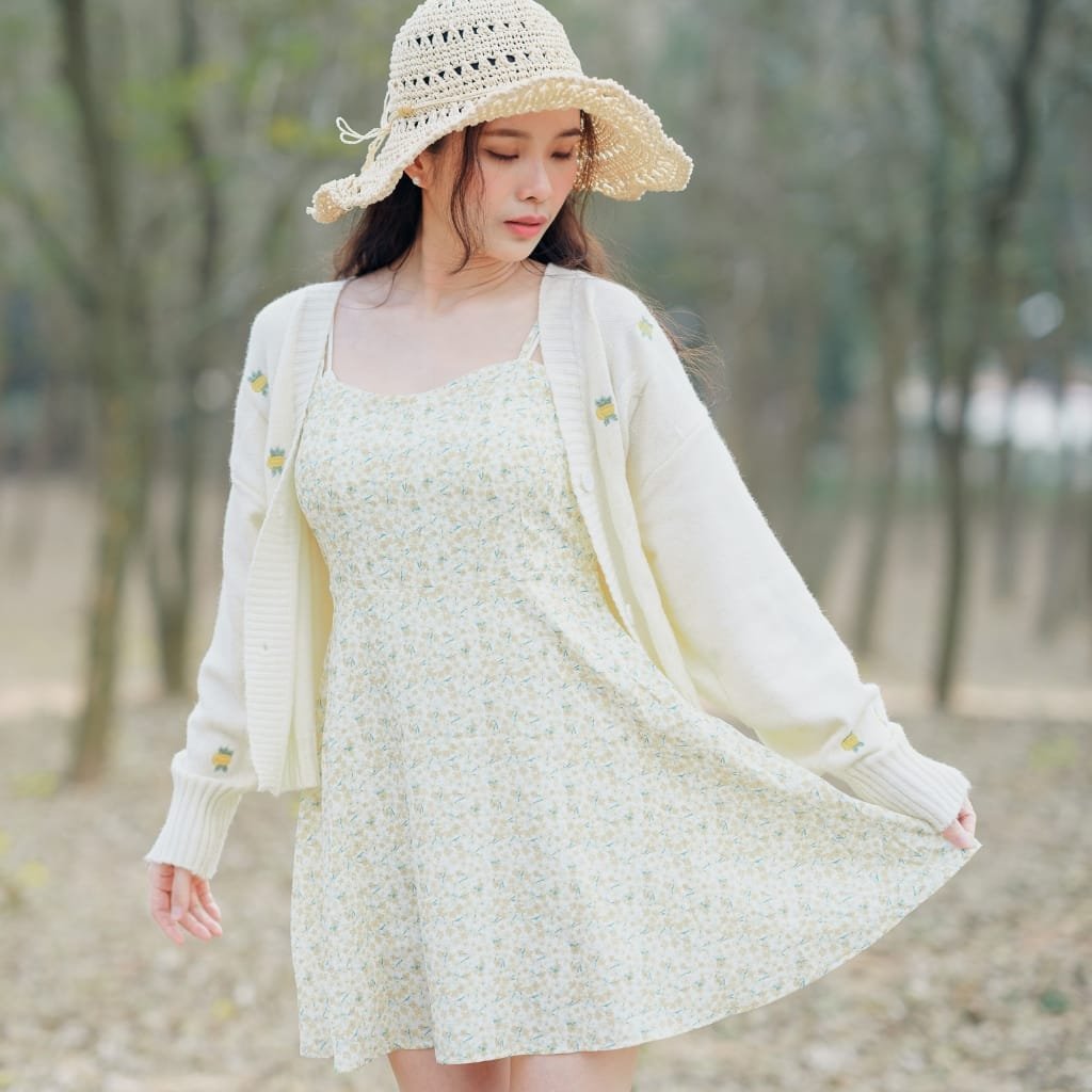 A young woman wearing a light-colored floral dress, a white cardigan with little embroidered details, and a wide-brimmed straw hat, standing outdoors among trees. She is gently holding the hem of her dress, with a soft and peaceful expression.