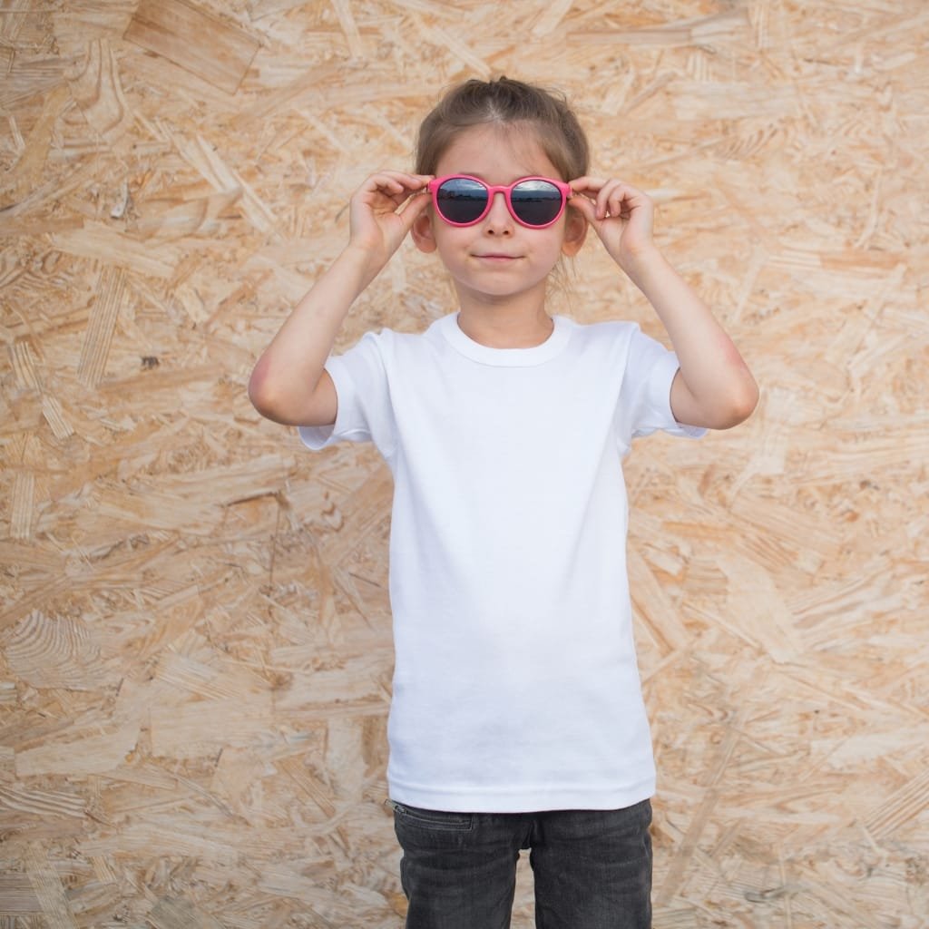 A young girl wearing a plain white cupro shirt and pink sunglasses, standing in front of a wooden wall.