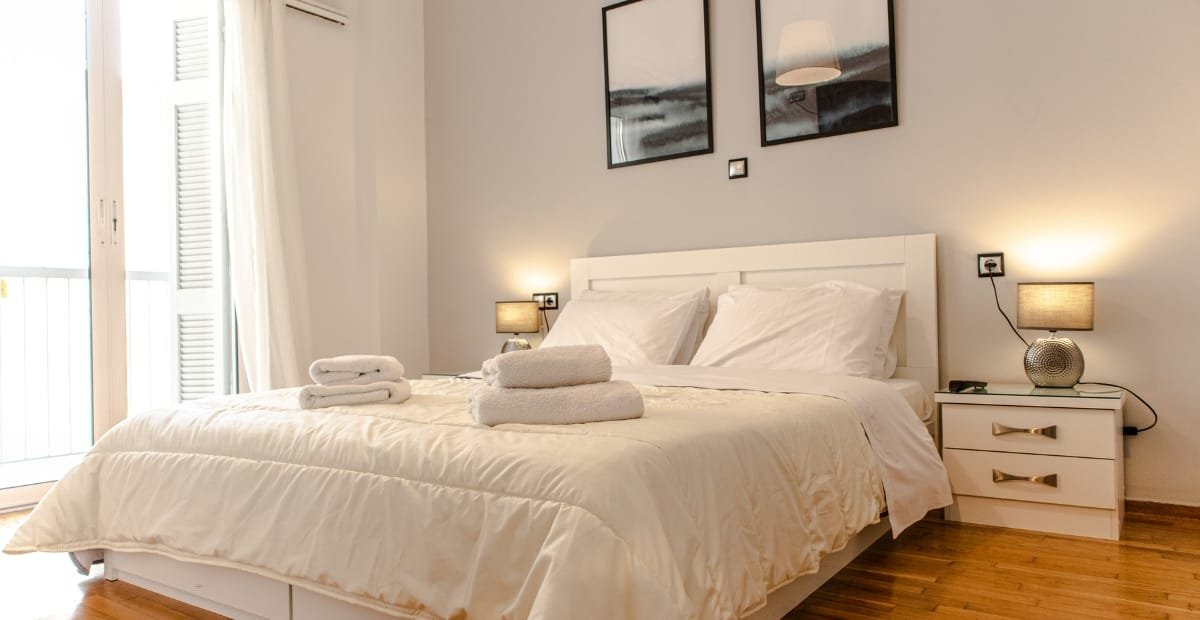 A neatly made bedroom featuring a white bed with folded towels, two bedside tables with lamps, and framed artwork above the headboard. There is a door leading to a balcony with closed shutters to the left.