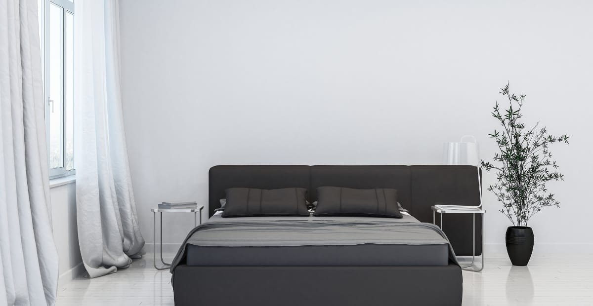 A modern bedroom with black linen bedding, featuring a neatly made bed with two matching black pillows, positioned against a white wall. There are two small side tables with books, a large black vase with a tall green plant, and white curtains on a window to the left.