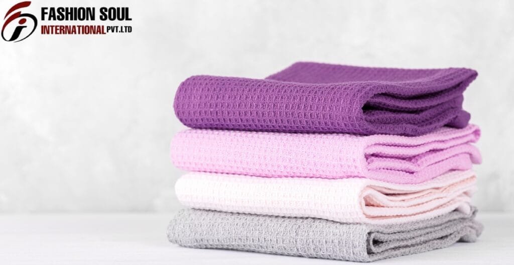 A stack of four folded waffle-knit towels in purple, pink, white, and gray, placed on a white surface against a light background with a "FASHION SOUL INTERNATIONAL PVT. LTD" logo in the top-left corner.