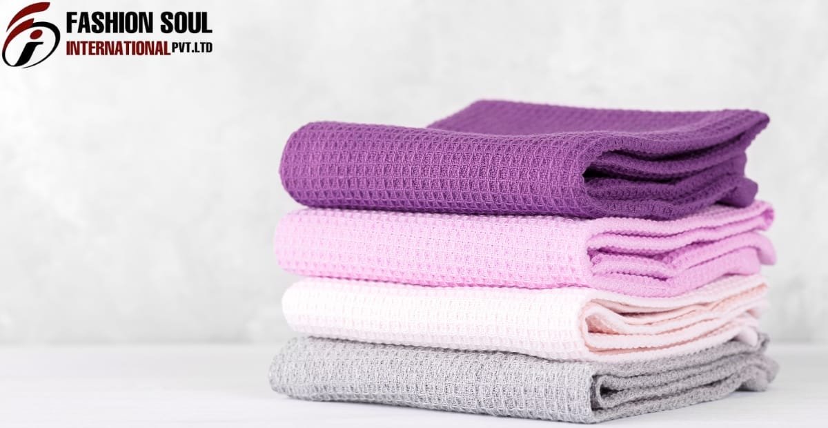 A stack of four folded waffle-knit towels in purple, pink, white, and gray, placed on a white surface against a light background with a "FASHION SOUL INTERNATIONAL PVT. LTD" logo in the top-left corner.