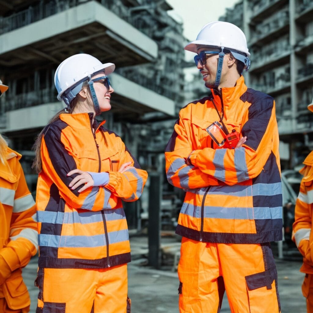 Two construction workers, a woman and a man, are standing outdoors at a construction site, wearing bright orange safety jackets, white hard hats, and safety glasses. They are smiling and engaging in a conversation with arms crossed, with a partially constructed building in the background.