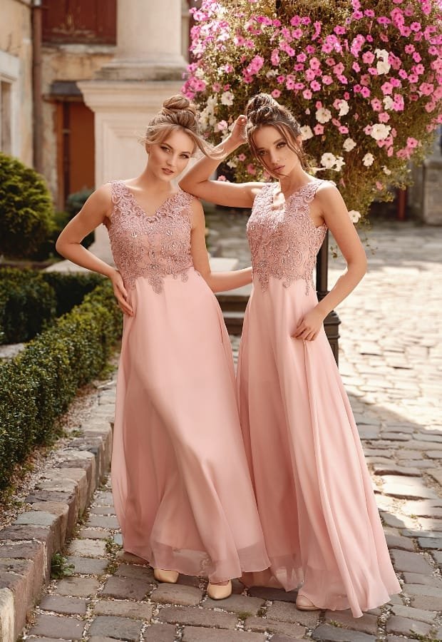 Two women wearing elegant pink ball gown dresses with lace embroidery on the bodice, standing outdoors on a cobblestone path surrounded by greenery and blooming flowers.