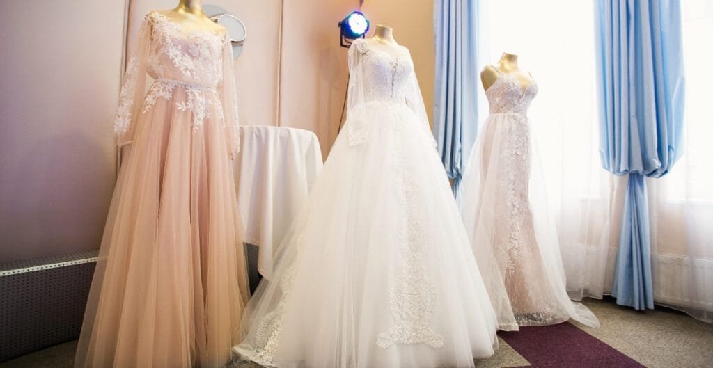 Four elegant ball gowns displayed on mannequins, each with intricate lace and tulle details, set against a softly lit background with blue curtains.