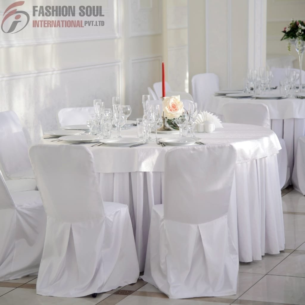 A round banquet table set for a formal event, covered with a white tablecloth and surrounded by white chairs with matching covers and tied with brown sashes. The table is elegantly decorated with wine glasses, plates, cutlery, a pink rose centerpiece, a tall red candle, and some decorative elements. The background shows a softly lit, white-walled room with elegant paneling.