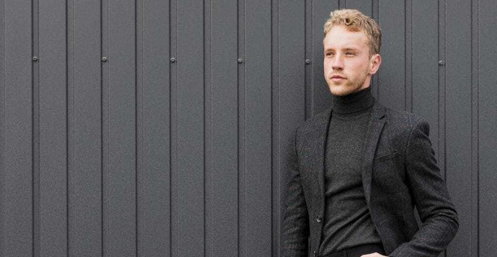 A man wearing a black sport coat and a turtleneck sweater, standing against a dark grey metal wall.