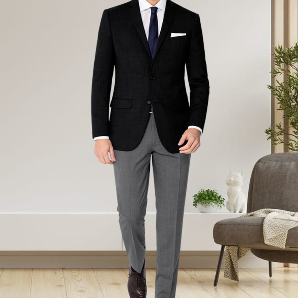 A man dressed in a formal black sport coat paired with light grey dress pants, white shirt, navy tie, and dark shoes, standing in a modern indoor setting.