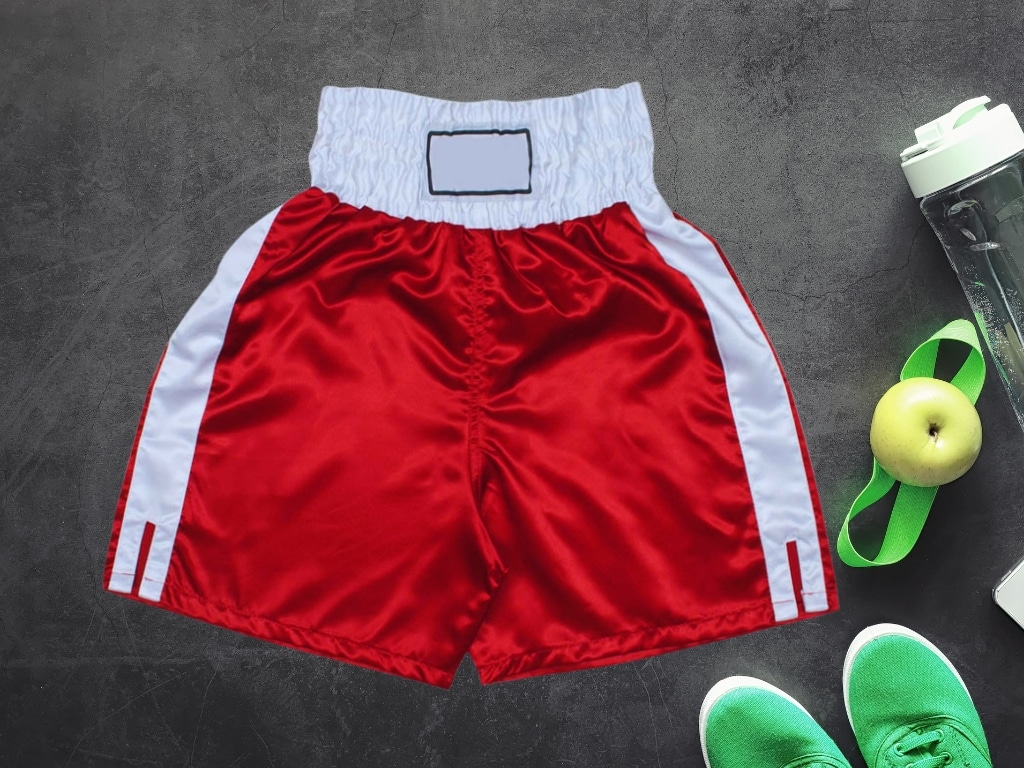 Red and white boxing shorts with a wide elastic waistband, laid flat on a dark surface alongside a water bottle, green apple, green shoes, and a green fitness band.