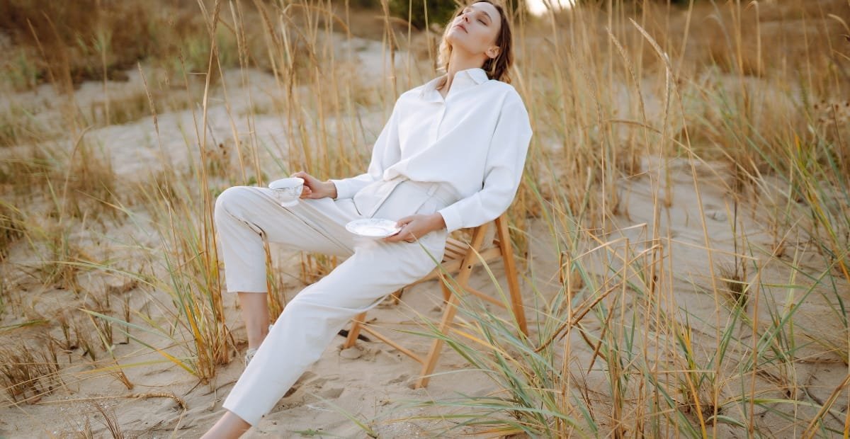 A woman dressed in white sits on a wooden chair on a sandy beach surrounded by tall, dry grass. She has her eyes closed and is holding two plates, looking relaxed and serene.