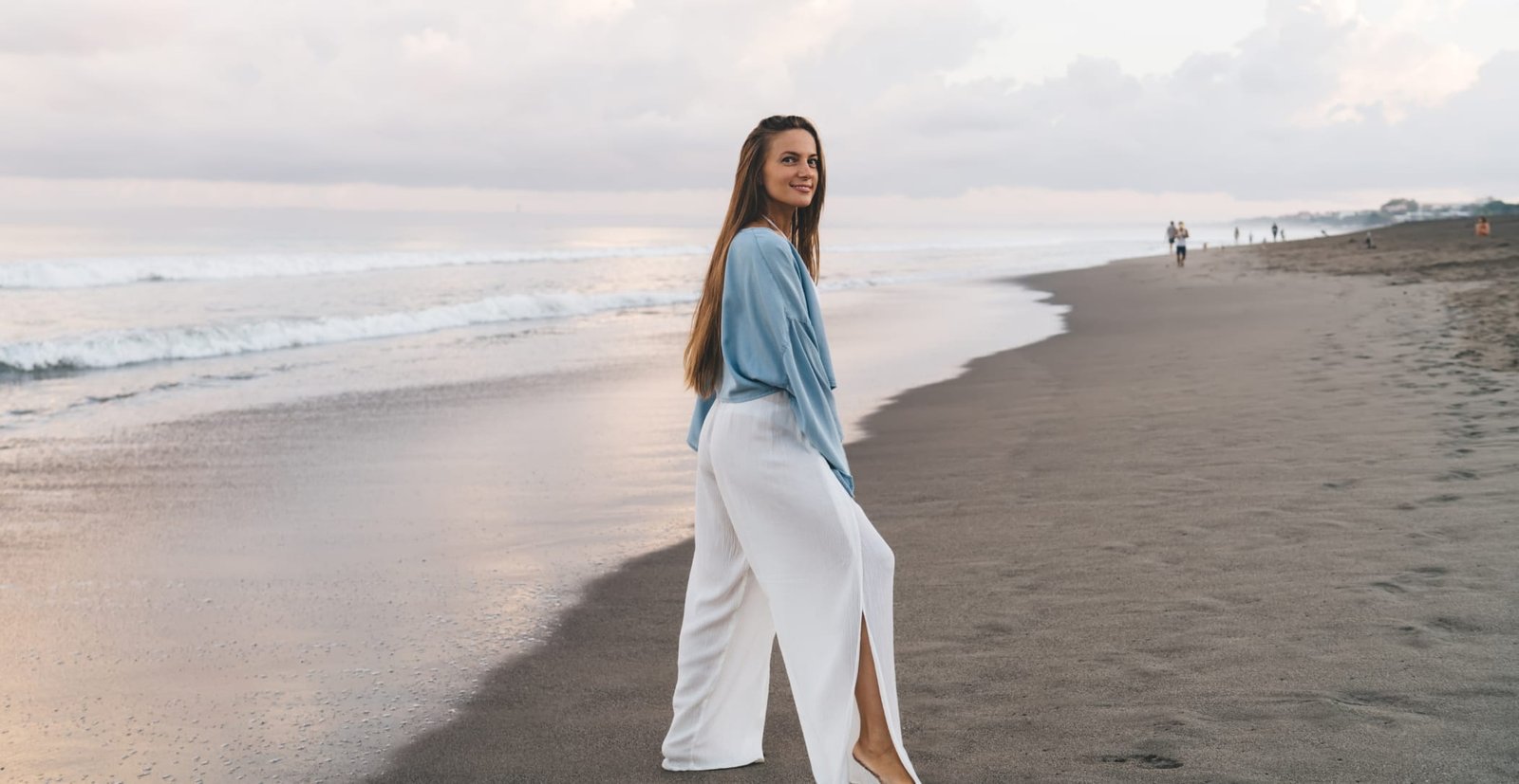 A woman standing on a sandy beach near the water, smiling and looking at the camera. She is wearing a light blue top and white wide-leg pants, with waves and a cloudy sky in the background.