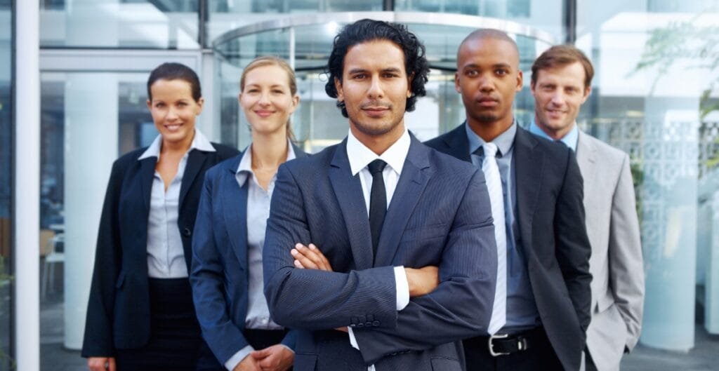 A group of five professionally dressed businesspeople standing outdoors in front of a modern office building, with a man in the foreground crossing his arms confidently.
