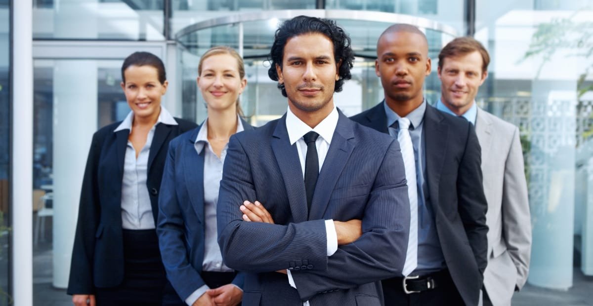 A group of five professionally dressed businesspeople standing outdoors in front of a modern office building, with a man in the foreground crossing his arms confidently.