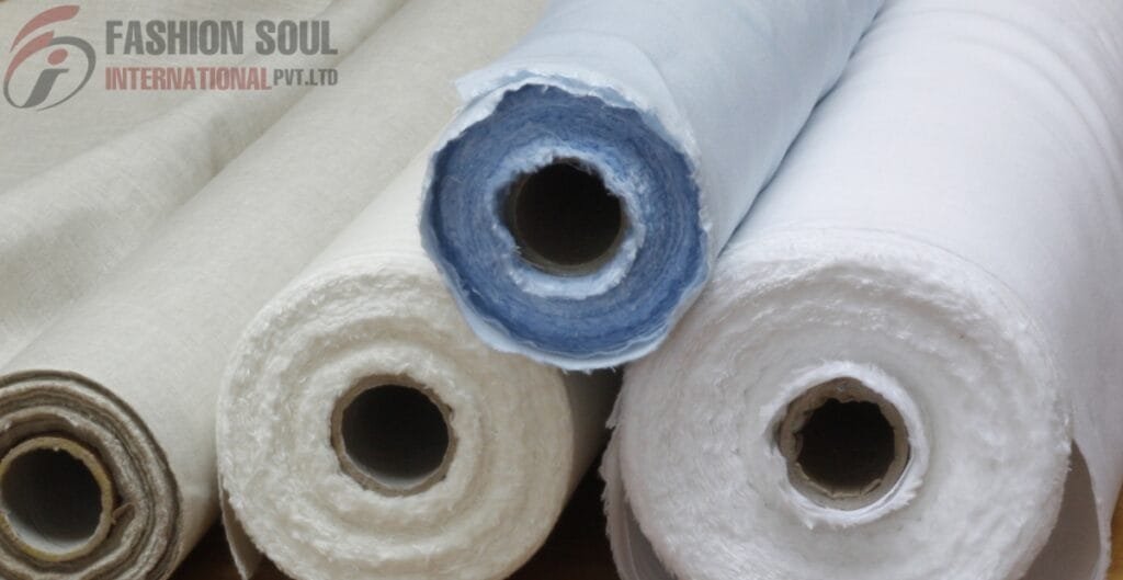 Close-up of multiple rolls of white and beige fabric or textile material wound around cardboard tubes, with one roll featuring a light blue edge. The rolls are piled together, and there's a logo in the top left corner that reads "FASHION SOUL INTERNATIONAL PVT. LTD."