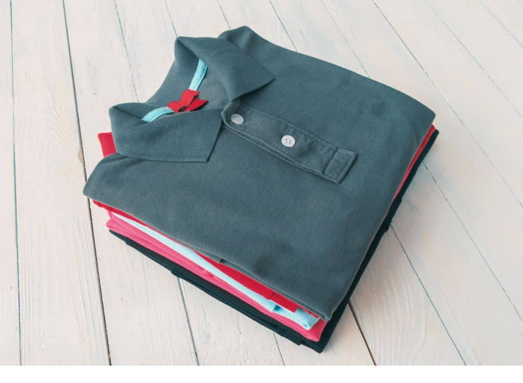 Folded stack of polo shirts in multiple colors, with the top shirt being a gray polo with a white label, placed on a light wooden surface.