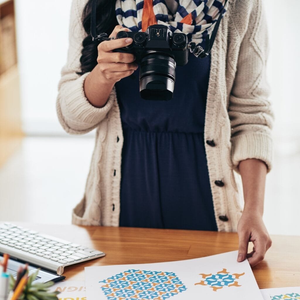  A person wearing a beige cardigan and scarf is holding a camera, with one hand resting on a table that has colourful artwork and a keyboard on it.