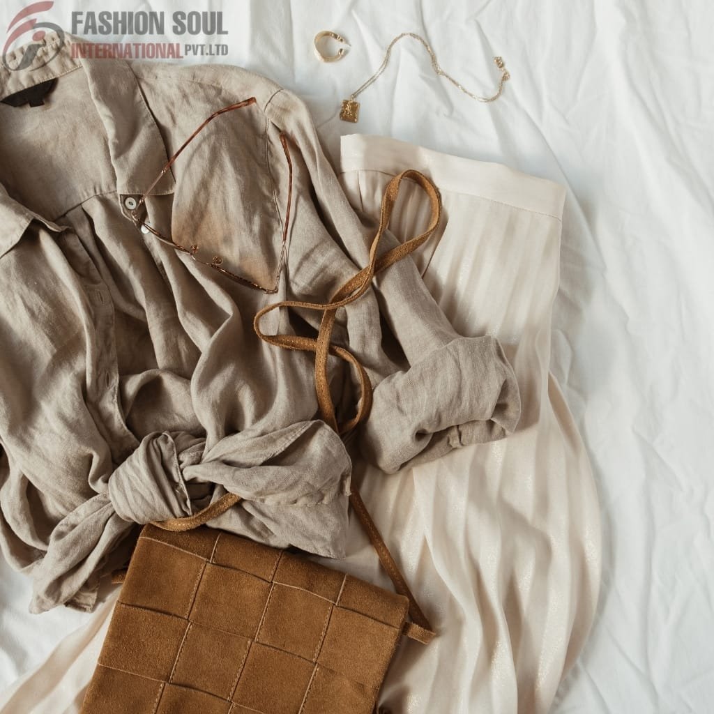 Flat lay of fashionable clothing and accessories on a white fabric background, including a beige blouse, cream-colored skirt, brown suede handbag, jewelry, and glasses.