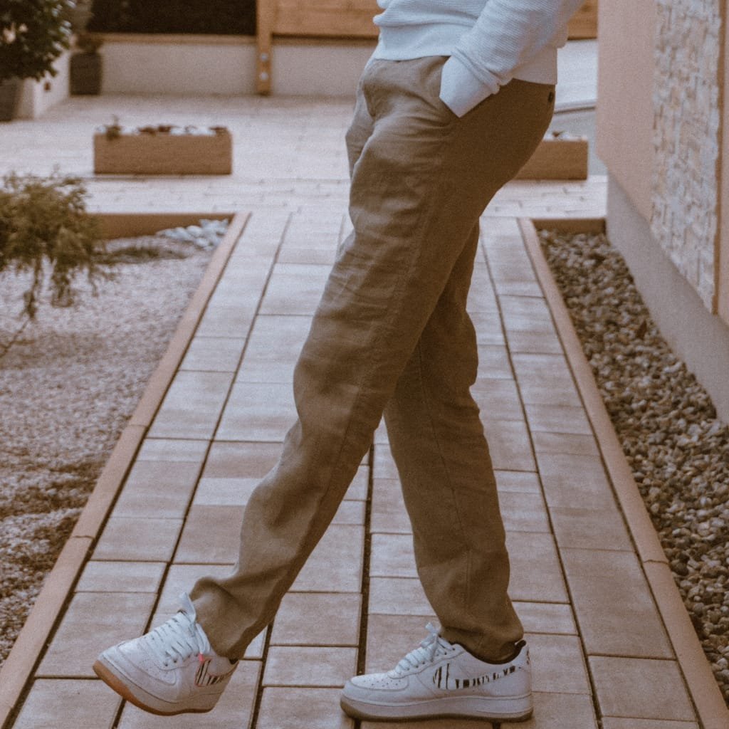 A person standing on a wooden pathway outdoors, wearing beige pants, white sneakers with black stripes, and a light gray hoodie with hands in pockets. The face is not visible.
