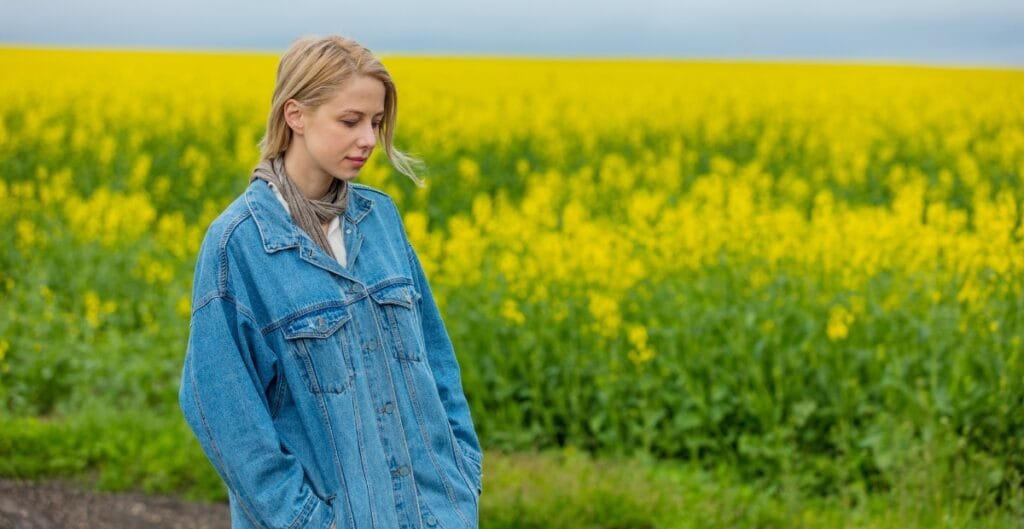A young woman with blonde hair, wearing a denim jacket, stands in a field of yellow flowers with a contemplative expression.
