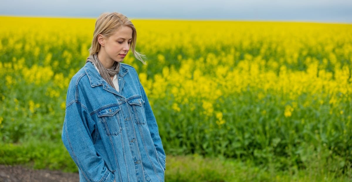 A young woman with blonde hair, wearing a denim jacket, stands in a field of yellow flowers with a contemplative expression.