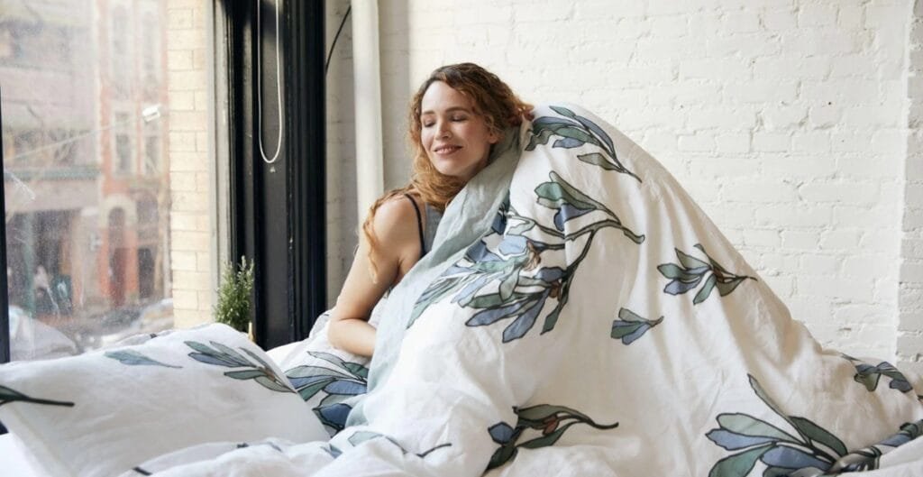 A woman with curly hair is sitting in bed near a large window, smiling and wrapped in a white blanket with a leafy pattern. She appears relaxed and content.