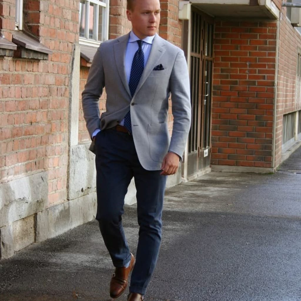A man walking along a city sidewalk wearing a smart outfit consisting of dark dress pants paired with a light gray tailored navy sport coat, a blue dress shirt, a patterned navy tie, and brown dress shoes.