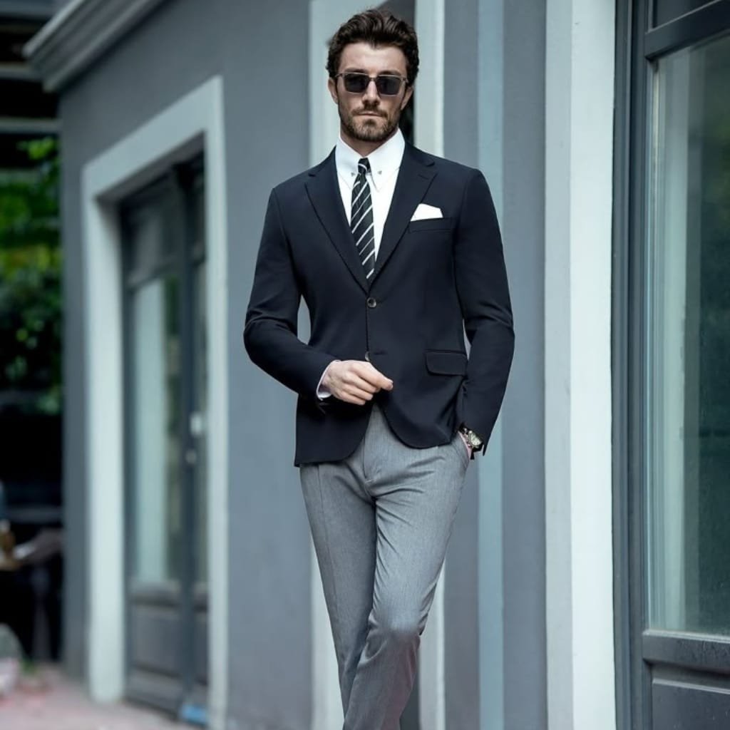 A stylish man wearing a black sport coat paired with a white shirt, gray trousers, and sunglasses, standing casually outside a modern building.