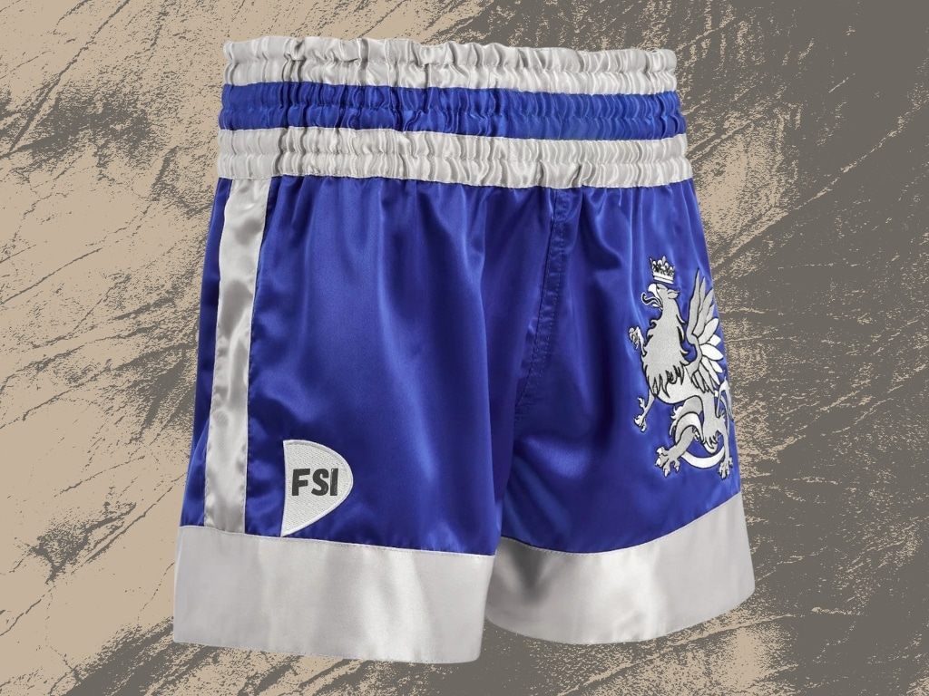 Blue and white satin boxing shorts with a crest and "FSI" logo, laid flat on a textured surface.