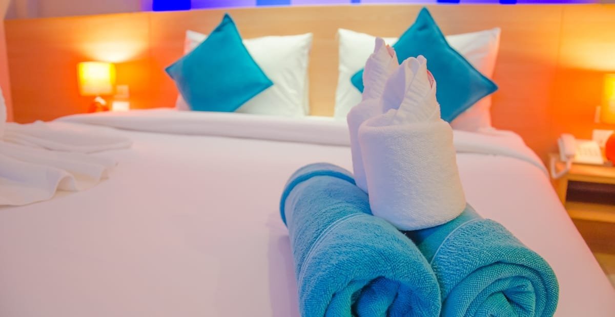 A neatly made hotel bed with two rolled towels shaped into swans in the foreground, four pillows (two white and two blue), and two warm-colored bedside lamps creating a cozy atmosphere.