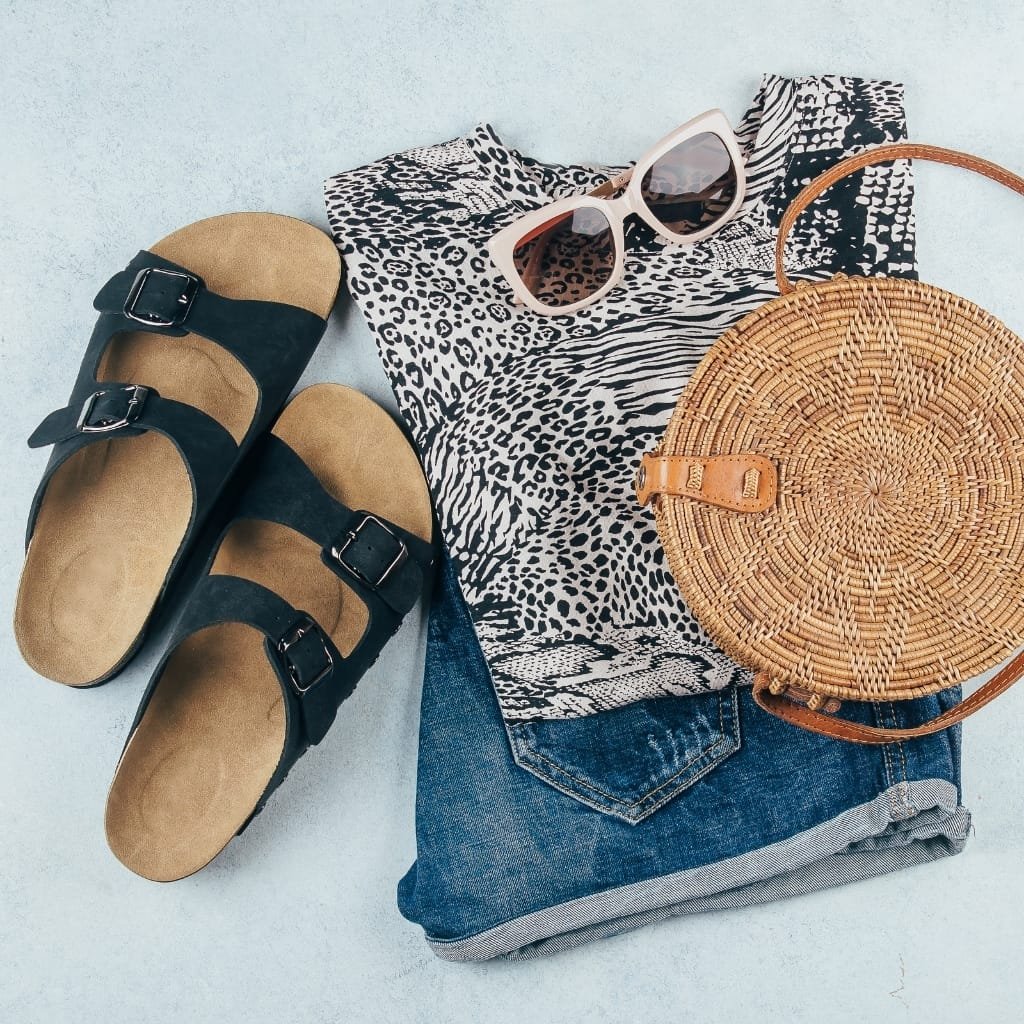 A flat lay of fashion accessories including a pair of black and tan sandals with buckles, a pair of white sunglasses, a black and white animal print fabric, a woven round handbag with a leather strap, and a pair of rolled-up denim shorts, all arranged on a light-colored surface.