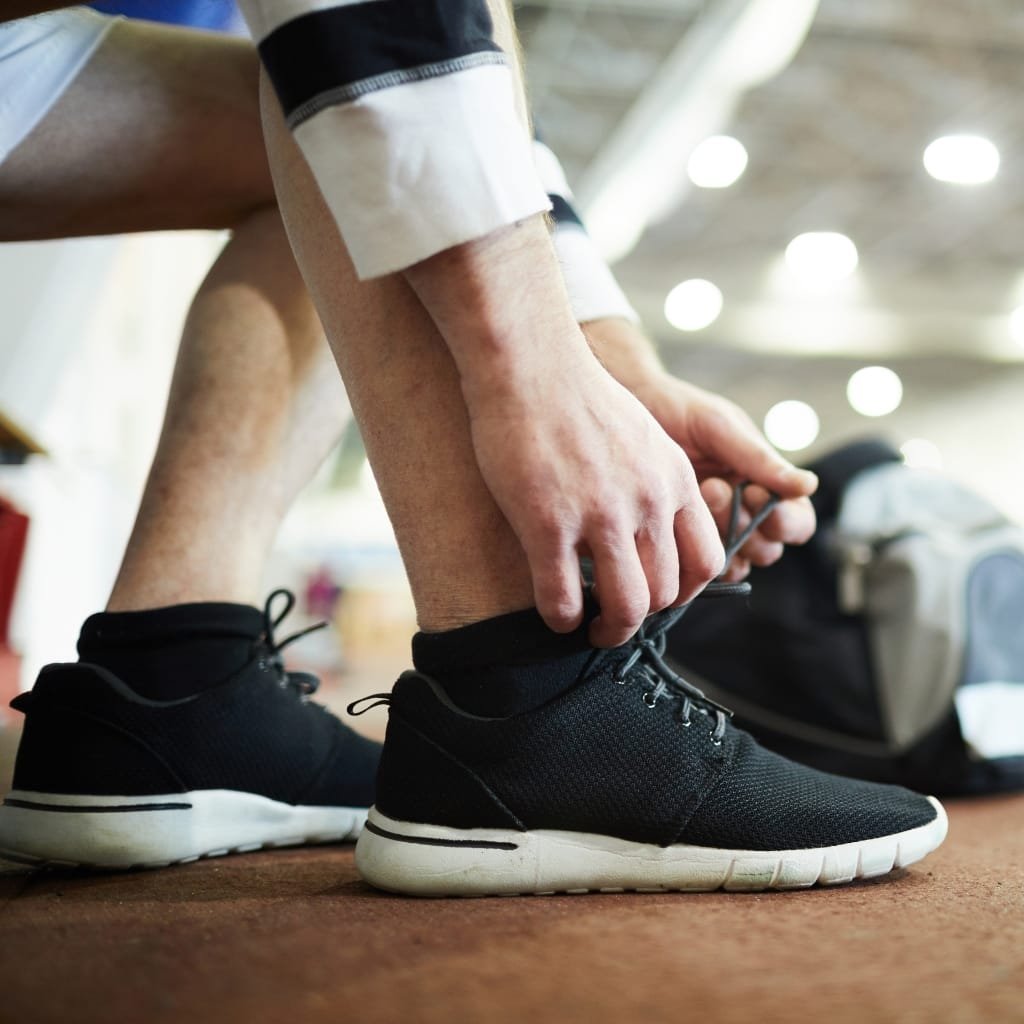 A person tying the laces on a black athletic sneaker, with a focus on their hands and shoes. The individual is wearing black socks and is seated indoors, possibly on a sports or gym floor.