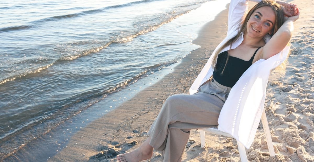 A woman with long hair smiling and relaxing on a beach, sitting on a chair with her arms behind her head, wearing a black top and beige pants, with the ocean and sand in the background.