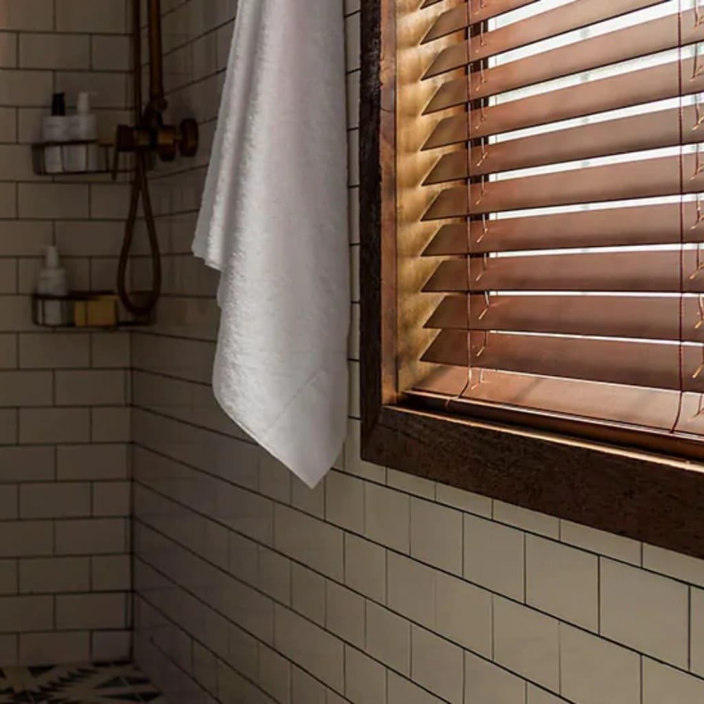 Close-up of a bathroom corner with beige tiled walls, a white towel hanging, and wooden window blinds.