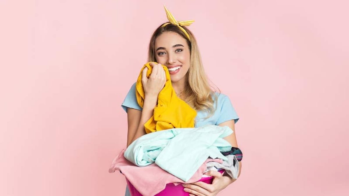 A smiling woman with a yellow headband holds a heap of colorful clothes against a pink background.